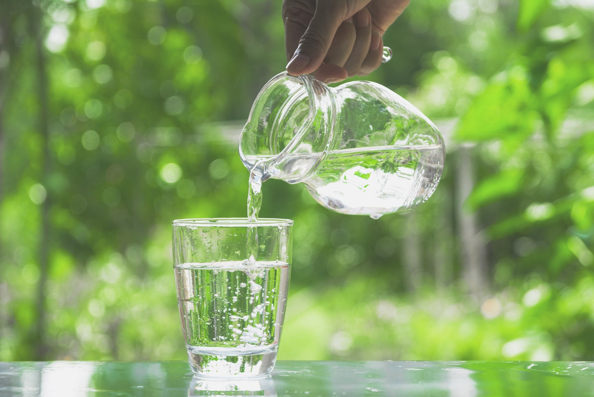Eine Hand gießt frisches, klares Wasser aus einem Glaskrug in ein Trinkglas. Im Hintergrund ist grüne Natur mit weichem Bokeh zu sehen, was die Szene ruhig und erfrischend wirken lässt.