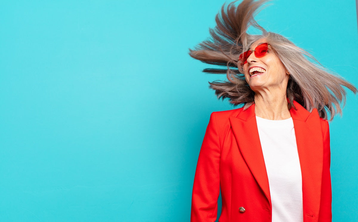 Energiegeladene Frau mit grauem Haar und rotem Blazer wirbelt lachend ihre Haare vor türkisblauem Hintergrund. Sie trägt eine rote Sonnenbrille und strahlt Lebensfreude und Selbstbewusstsein aus – Symbol für Vitalität und positives Lebensgefühl in der zweiten Lebenshälfte.