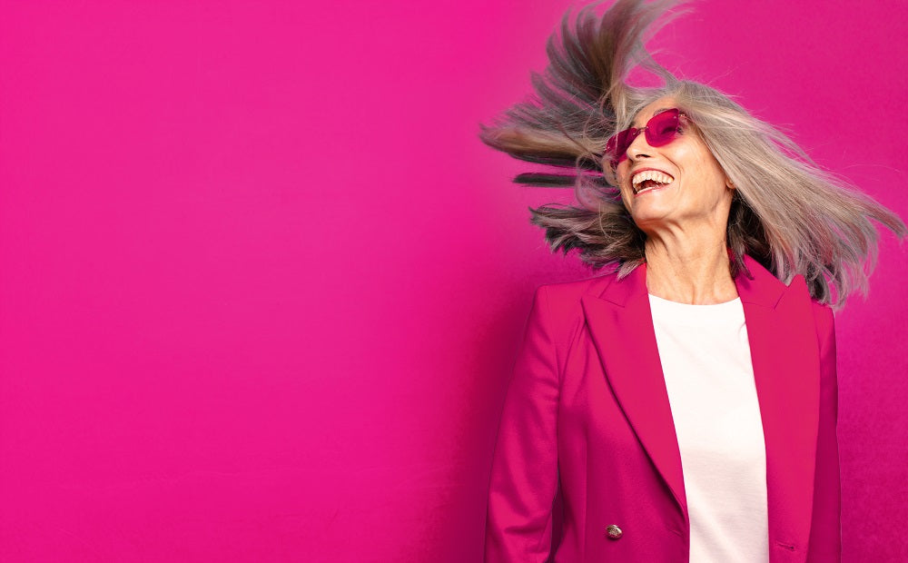 Frau mit grauen Haaren, pinker Sonnenbrille und pinken Blazer steht lachend vor pinken Hintergrund