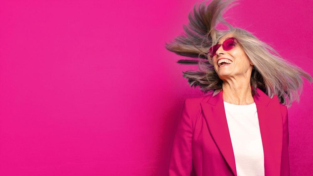 Frau mit grauen Haaren und pinker Sonnenbrille mit fliegenden Haaren befindet sich in den Wechseljahren.