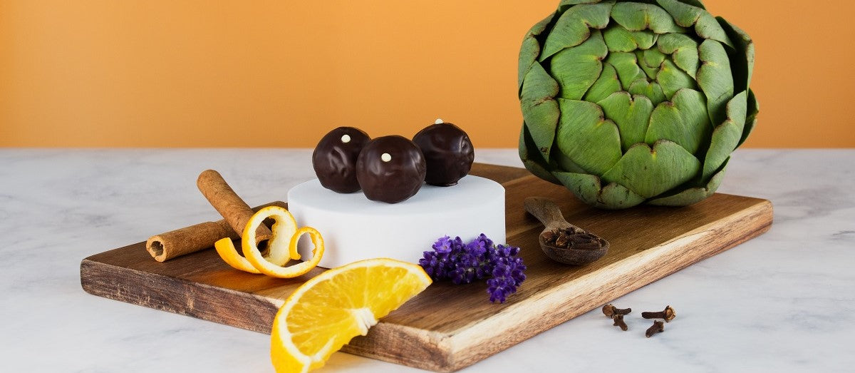 Bitterstern Schokoladentrüffel auf einem Holzbrett mit natürlichen Bitterstoffen: Artischocke, Zimtstangen, Orangenscheibe, Nelken, Lavendel und Zitronenzeste – edle Kombination aus bitteren Kräutern und hochwertiger Schokolade für Genuss und Wohlbefinden.