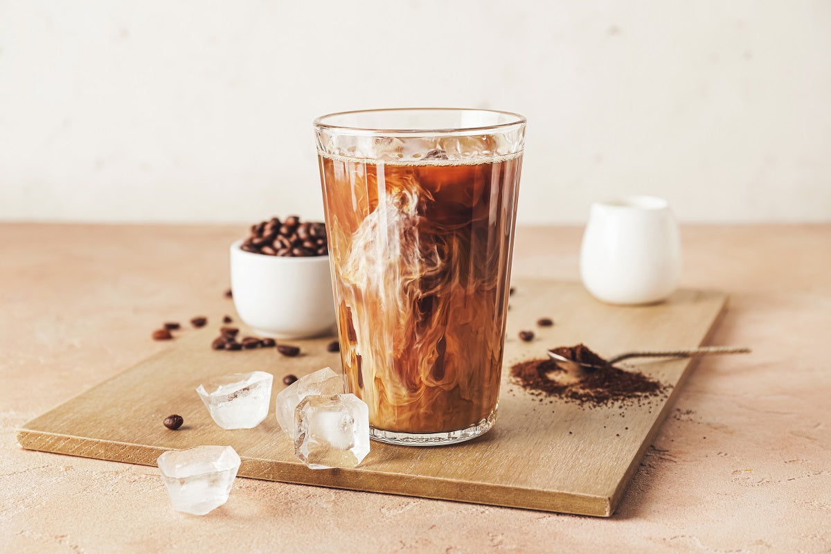 Ein erfrischender Eiskaffee mit einem Schuss Milch, serviert in einem hohen Glas mit Eiswürfeln. Im Hintergrund sind Kaffeebohnen, Kaffeepulver und eine kleine Milchkanne zu sehen – ideal kombiniert mit Bitterstern Tropfen für eine bittere Note und bessere Verträglichkeit.