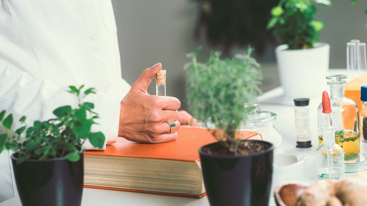 Laetitia Experten mit Reagenzglas in der Hand, Arztkittel, auf dem Tisch Heilkräuter und ein Buch