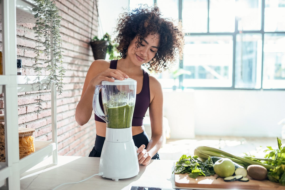 Junge Frau in Sportkleidung bedient einen Standmixer mit grünem Smoothie in einer hellen Küche. Auf der Arbeitsfläche liegen frisches Gemüse und Obst.