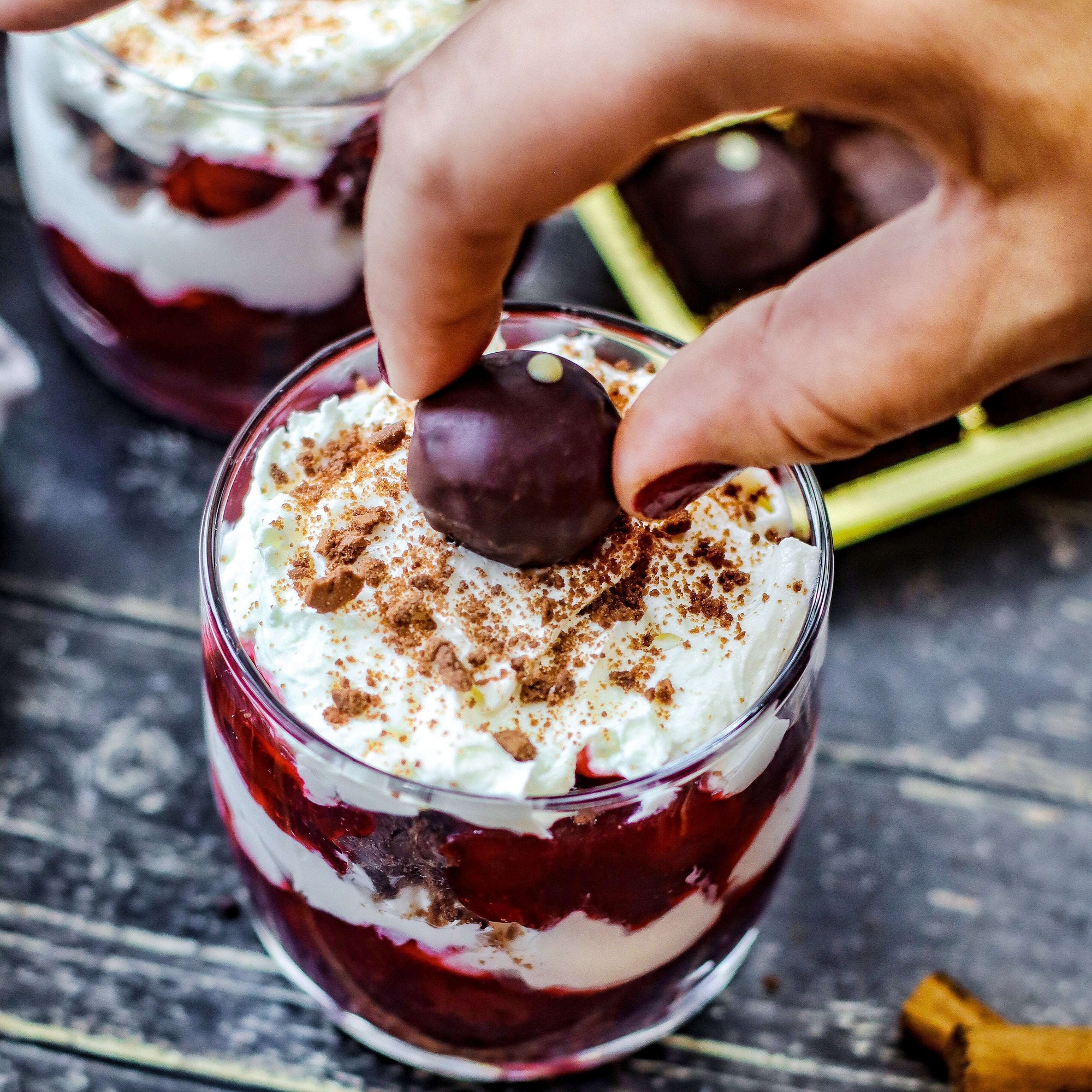 Ein Glas mit geschichteter Kirschdessert, Sahne und roten Früchten wird mit einem Bitterstern Schokoladentrüffel garniert, im Hintergrund eine offene Pralinenverpackung auf einem dunklen Holztisch.