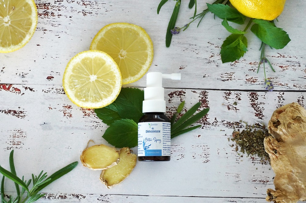 Bitterstern Bitter-Spray ohne Alkohol mit weißem Sprühaufsatz auf rustikalem, weiß gestrichenem Holzhintergrund. Umgeben von frischen Zutaten wie Zitronenscheiben, Ingwer, Minze, Lavendel, Rosmarin, Zitronenmelisse und getrockneten Kräutern.