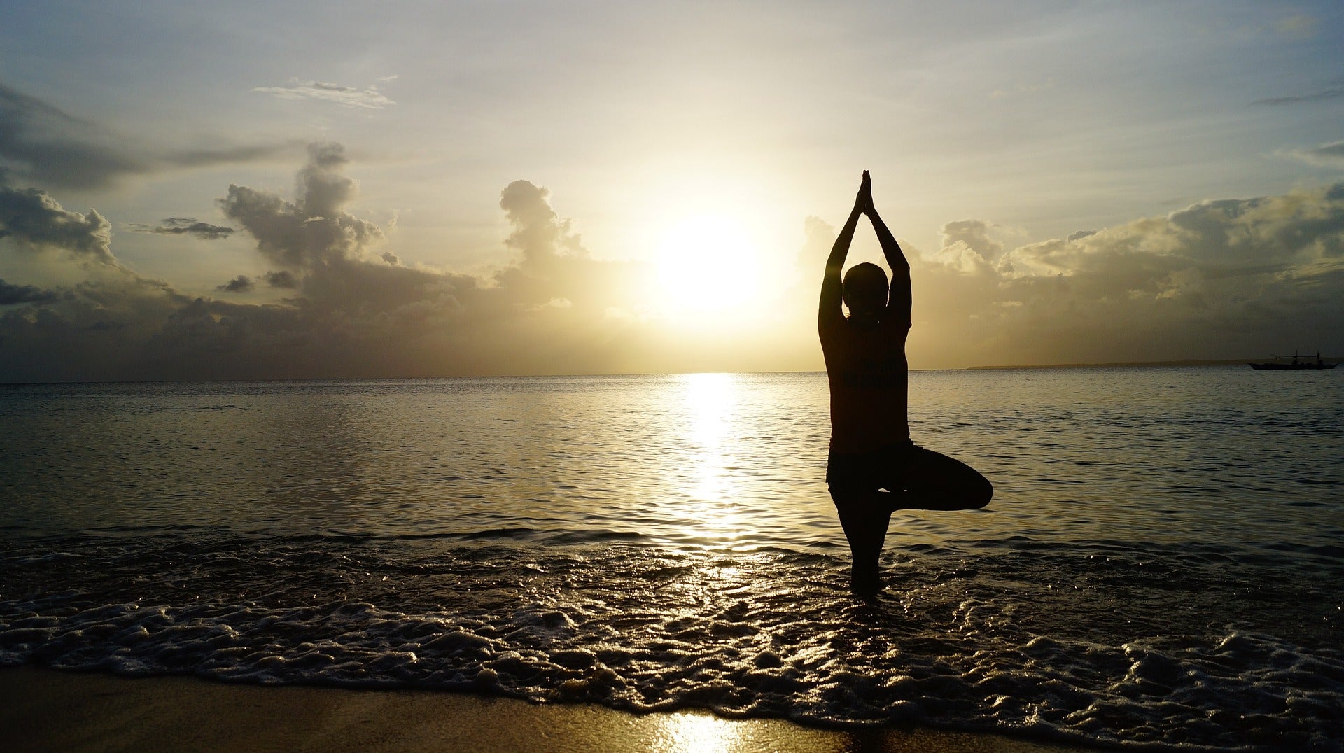 Person im Sonnenuntergang bei Yoga am Meer, stehend in der Baumhaltung im seichten Wasser vor ruhigem Horizont – Symbol für innere Balance und Gelassenheit.