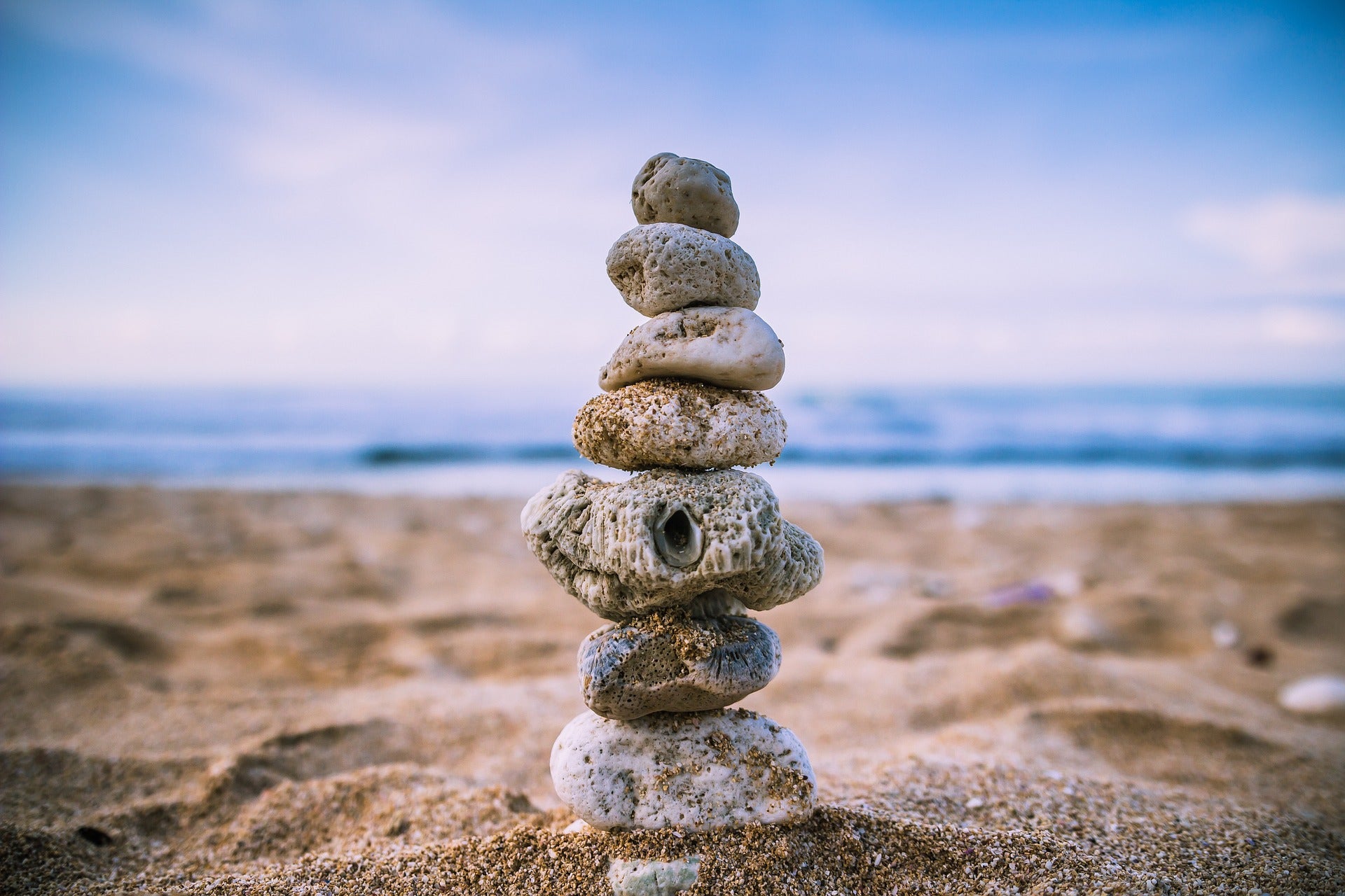 Aus kleinen Steinen aufgeschichteter Steinturm im Sand am Strand, mit ruhigem Meer und blauem Himmel im Hintergrund – Symbol für Balance und Achtsamkeit.