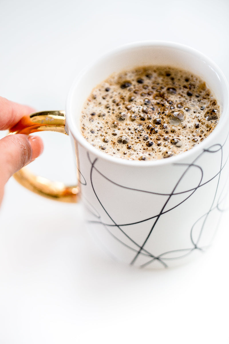 Kaffee in einer weißen Tasse mit schwarzen Ornamenten und goldenem Henkel