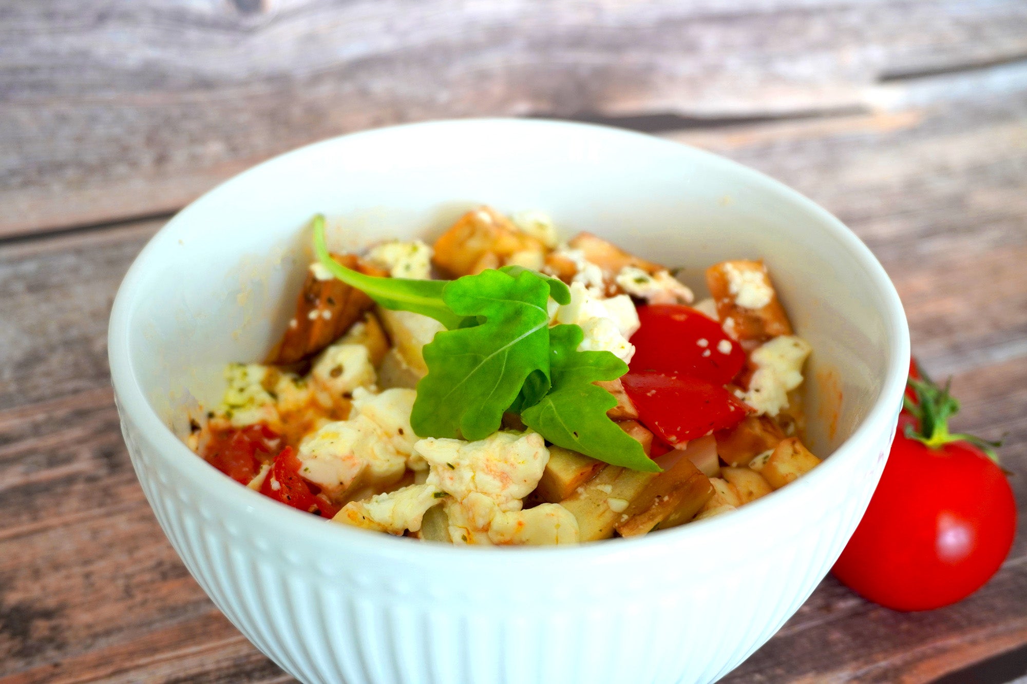Weiße Schüssel mit einem Gemüsepäckchen aus gebratenem Gemüse, Fetakäse und frischem Rucola. Daneben liegt eine rote Tomate auf einem Holztisch.