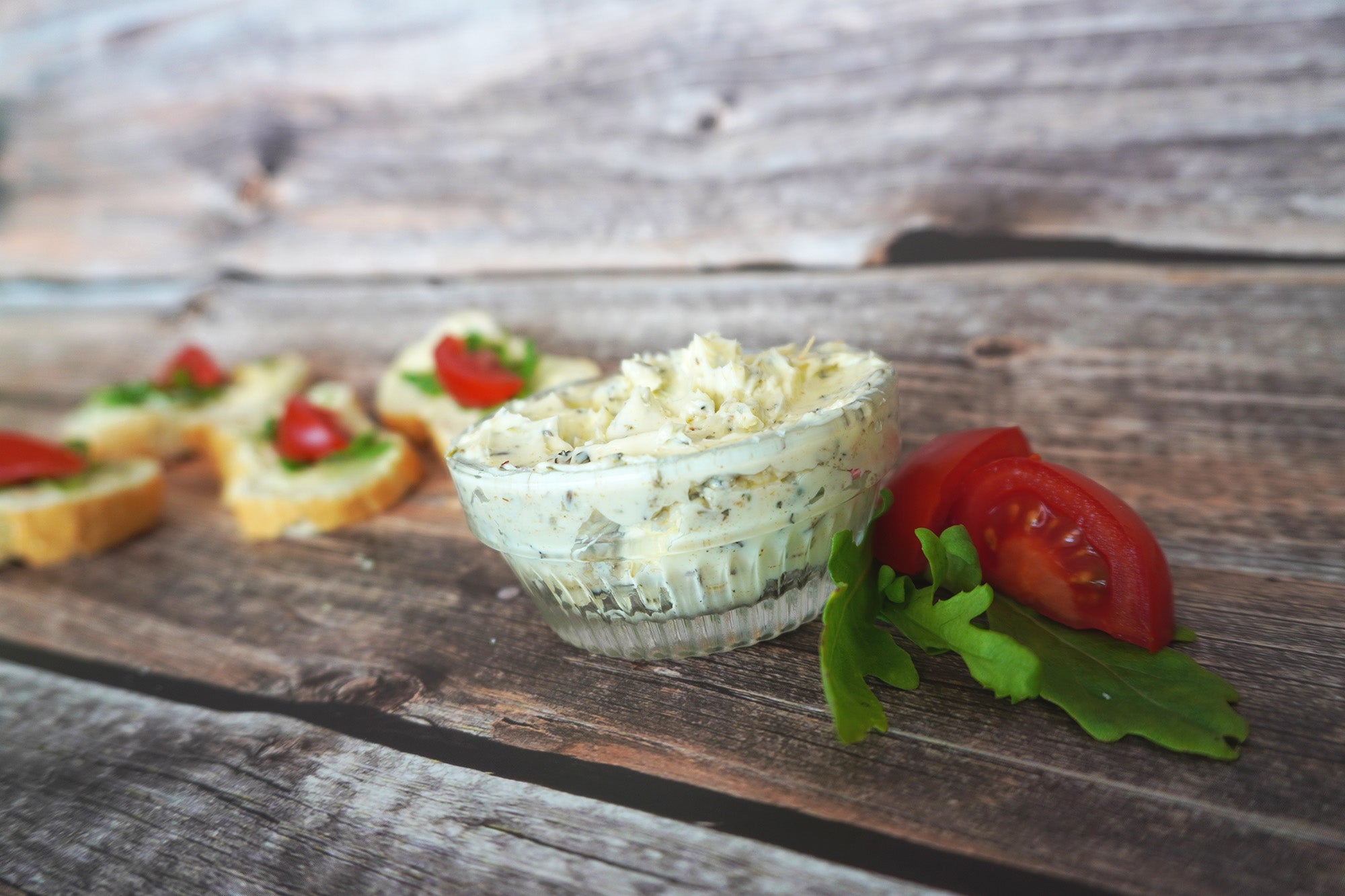 Selbstgemachte Kräuterbutter in einer kleinen Glasschale, daneben frische Tomatenstücke und Rucolablätter auf rustikalem Holzuntergrund; im Hintergrund Baguettescheiben mit Aufstrich, Rucola und Tomate.