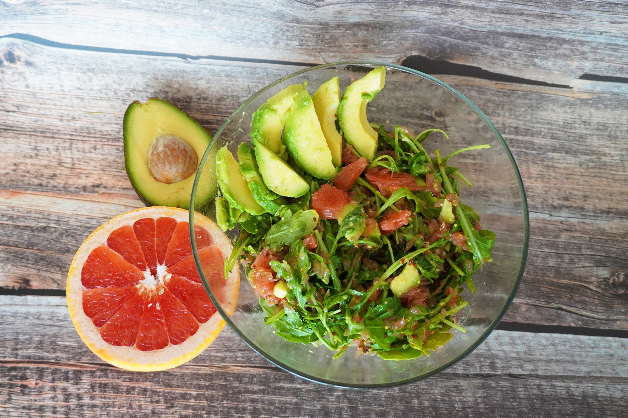 Frischer Rucolasalat mit Avocadostreifen und Grapefruitstücken in einer Glasschüssel, daneben eine halbierte Avocado und eine aufgeschnittene Grapefruit auf einem Holztisch.