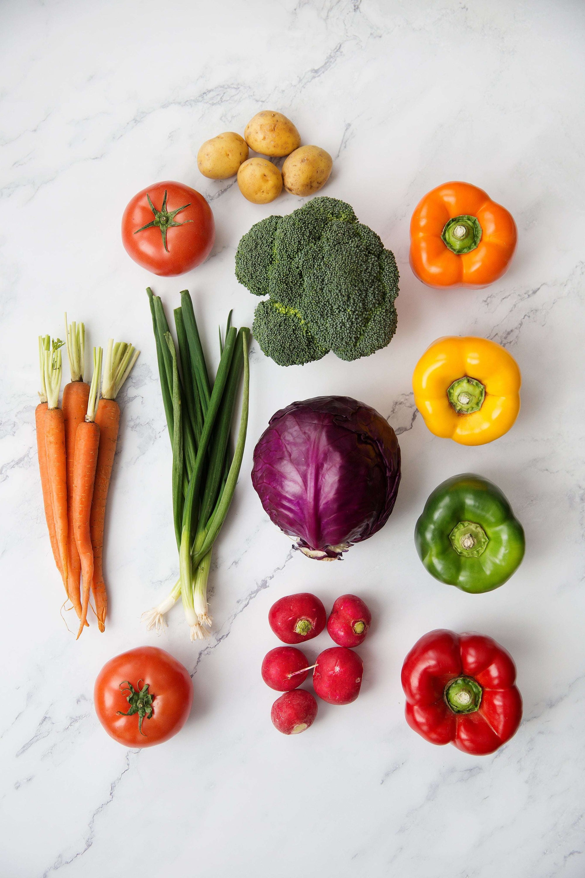 Ein bunter Mix aus frischem Gemüse – darunter Tomaten, Karotten, Frühlingszwiebeln, Paprika, Kartoffeln, Brokkoli, Rotkohl und Radieschen – liegt ordentlich arrangiert auf einer hellen Marmoroberfläche.