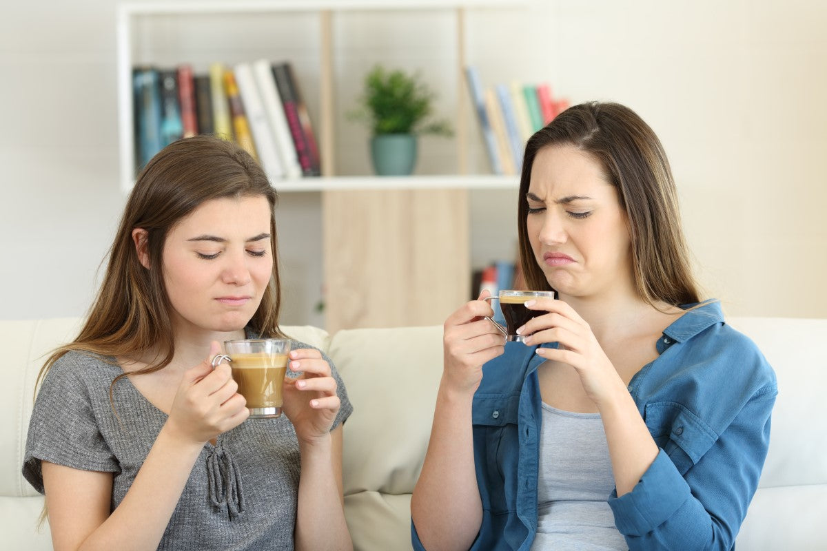 Zwei junge Frauen sitzen auf einem Sofa und halten jeweils eine Tasse Kaffee in der Hand. Beide schauen mit Ekel oder Unzufriedenheit auf ihre Getränke. Im Hintergrund ist ein Bücherregal mit Pflanzen und Büchern zu sehen.