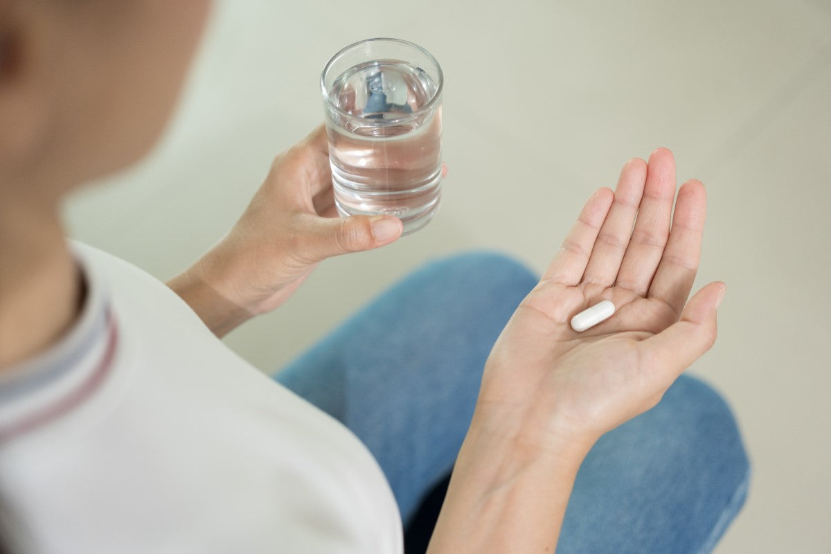Eine Frau hält in der linken Hand ein Glas Wasser und in der rechten Hand eine weiße Kapsel. Die Person trägt ein weißes Oberteil und eine blaue Jeans, die Szene ist aus der Vogelperspektive über die Schulter aufgenommen.