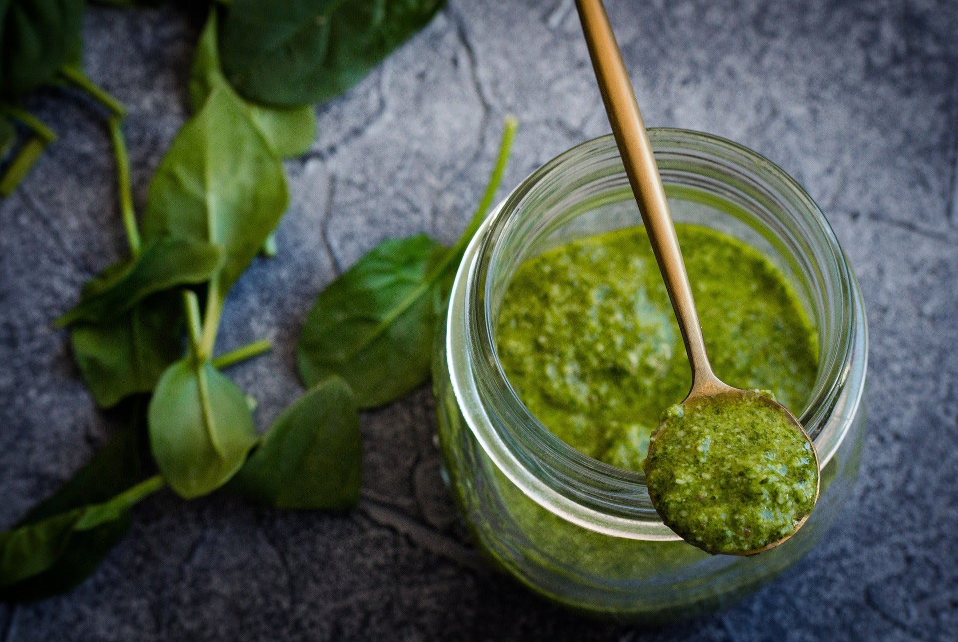 Glas mit grünem Pesto und goldener Löffel, umgeben von frischen Spinatblättern auf dunklem Steinuntergrund – würzig und natürlich.