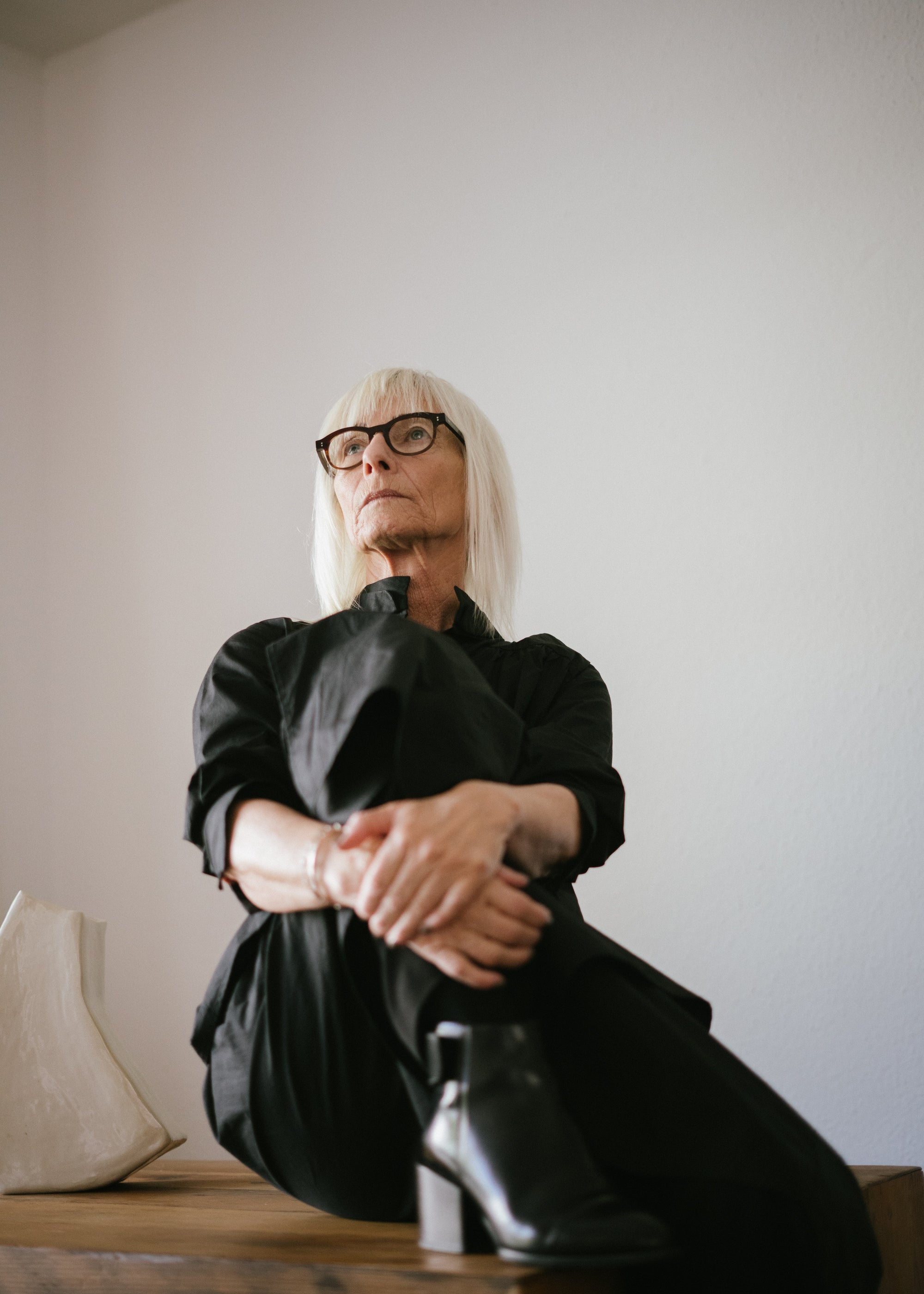 Ältere Frau mit weißblonden Haaren und Brille sitzt entspannt in schwarzem Outfit auf einem Holztisch vor heller Wand, modernes Ambiente, natürliche Ausstrahlung.