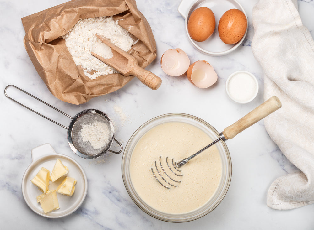 Backzutaten wie Mehl, Eier, Butter und Zucker liegen auf einer hellen Arbeitsfläche verteilt. In einer Schüssel befindet sich ein cremiger Teig mit Schneebesen – bereit zum Backen von Plätzchen nach Hildegard von Bingen.
