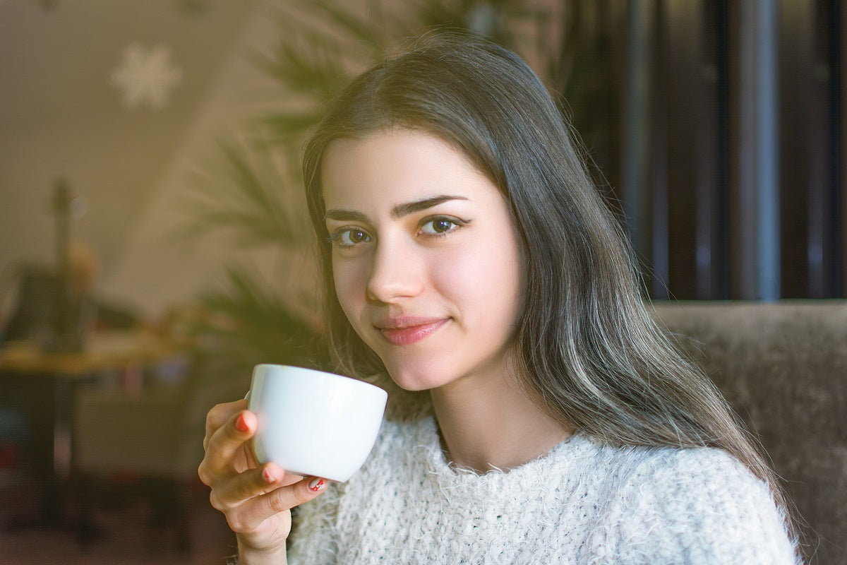 Junge Frau trinkt Kaffee aus einer weißen Tasse in einem Wohnzimmer mit einer Pflanze im Hintergrund