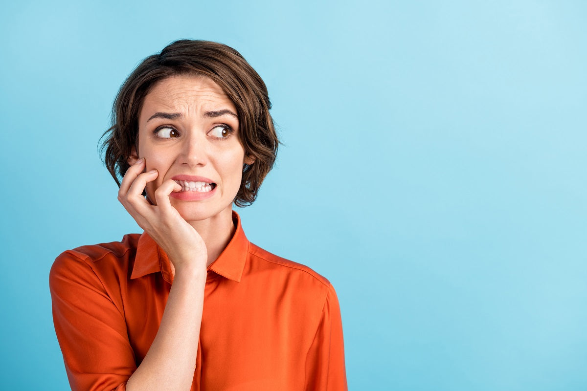 Junge Frau in orangefarbener Bluse blickt ängstlich zur Seite und beißt nervös auf ihre Fingernägel – Symbolbild für Unsicherheit, Nervosität oder Angst.