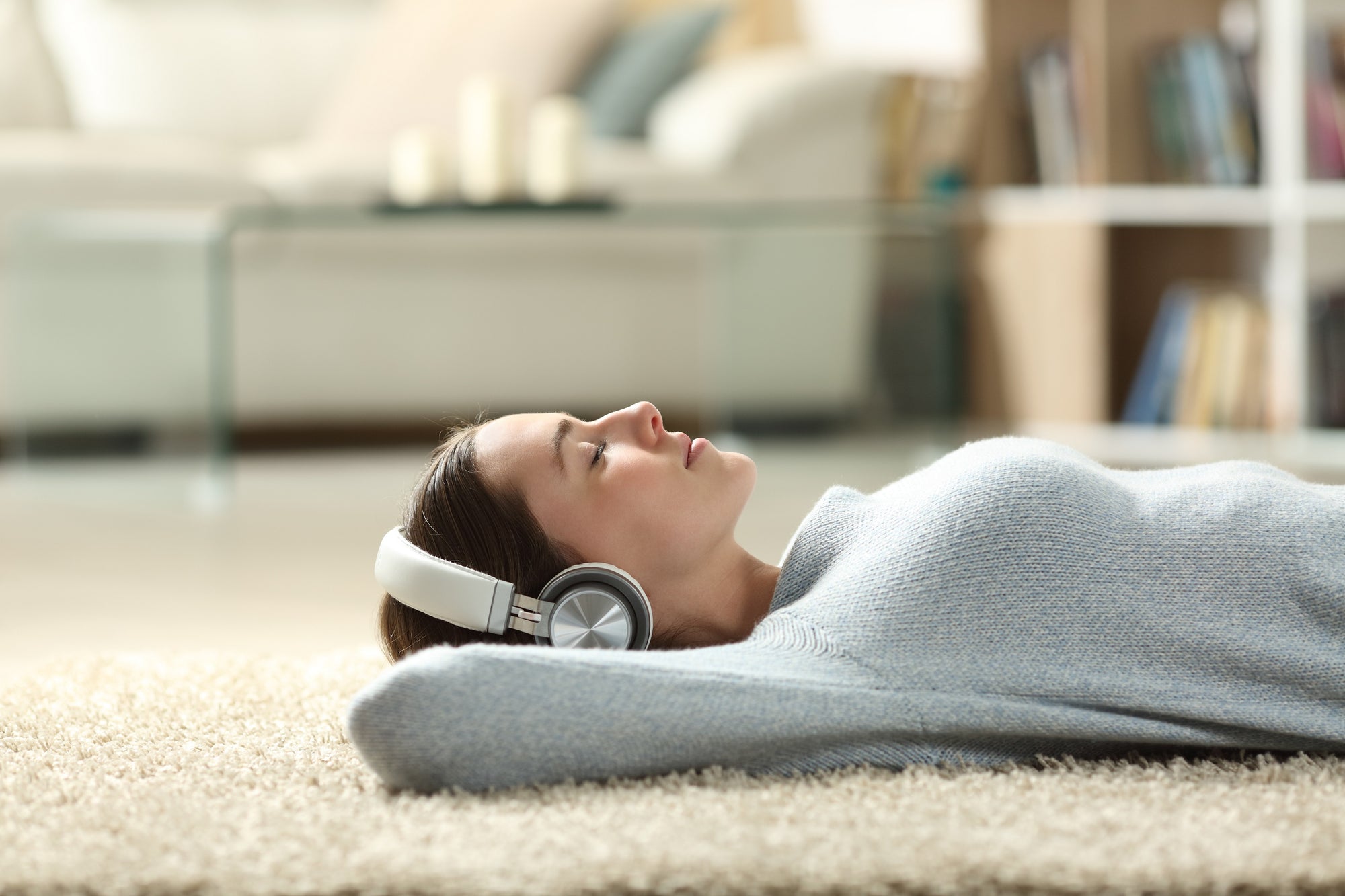Junge Frau liegt entspannt mit geschlossenen Augen auf einem weichen Teppichboden, trägt große Kopfhörer und hört Musik oder eine Meditation. Der Hintergrund zeigt ein gemütliches Wohnzimmer mit Sofa und Bücherregal.