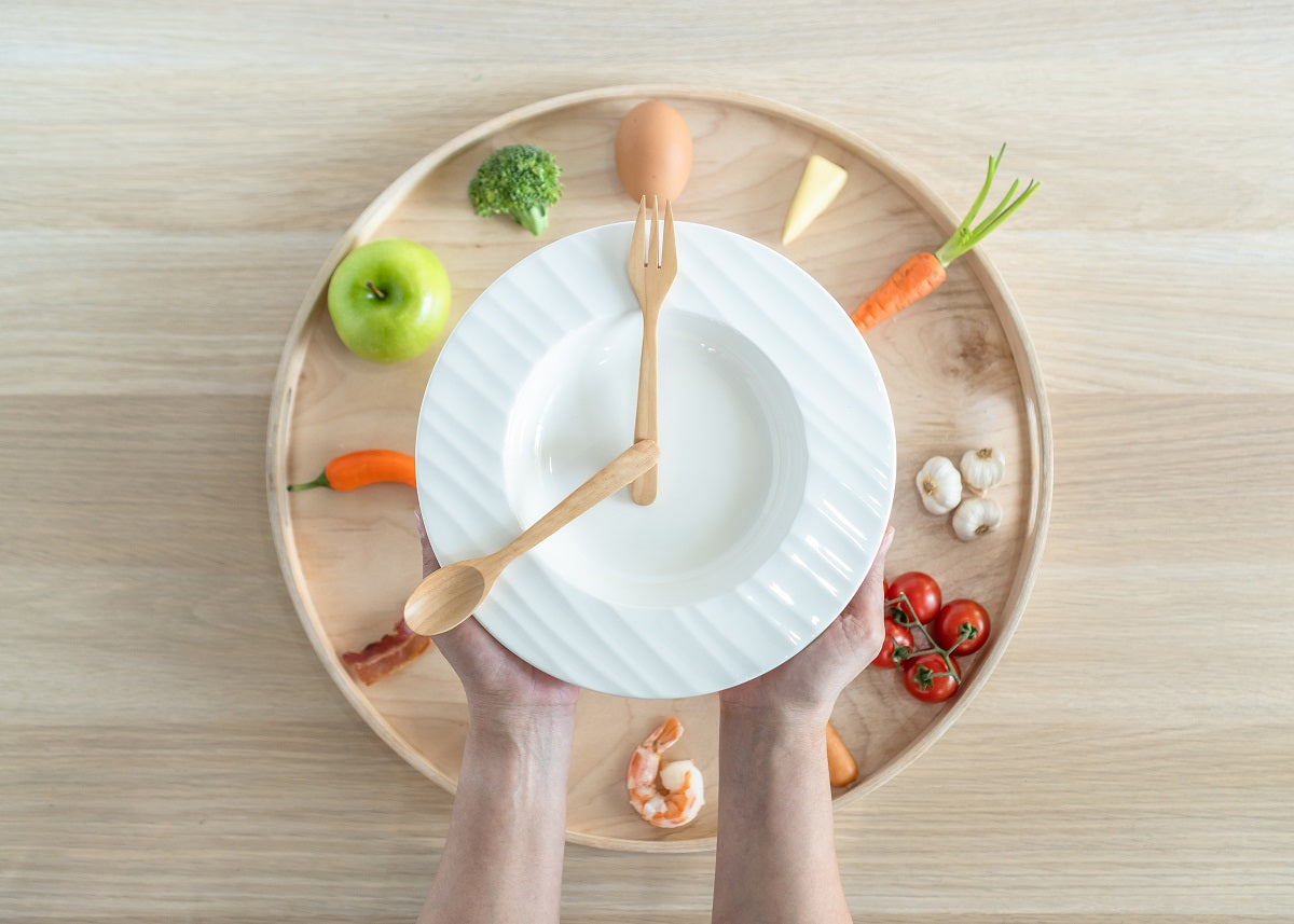 Zwei Hände halten einen weißen, leeren Teller mit Löffel und Gabel in Form eines Uhrzeigers auf einem runden Holztablett, das mit gesunden Lebensmitteln wie Apfel, Ei, Brokkoli, Karotten, Tomaten, Knoblauch, Käse und Garnelen dekoriert ist – Symbol für Intervallfasten, gesunde Ernährung und ausgewogene Mahlzeiten.