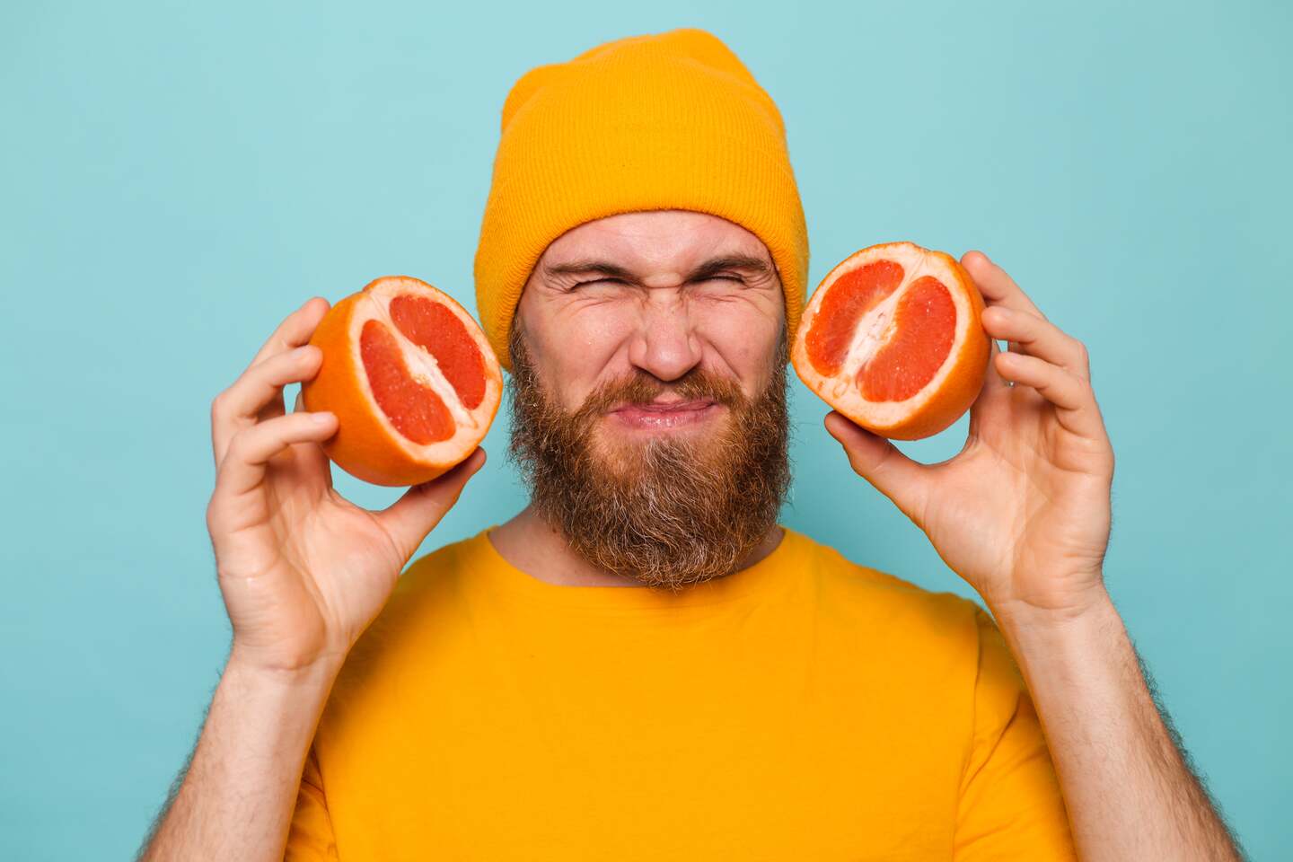 Mann mit Vollbart, gelber Mütze und gelbem Shirt hält zwei Hälften einer Grapefruit vor sein Gesicht und verzieht das Gesicht wegen des sauren Geschmacks