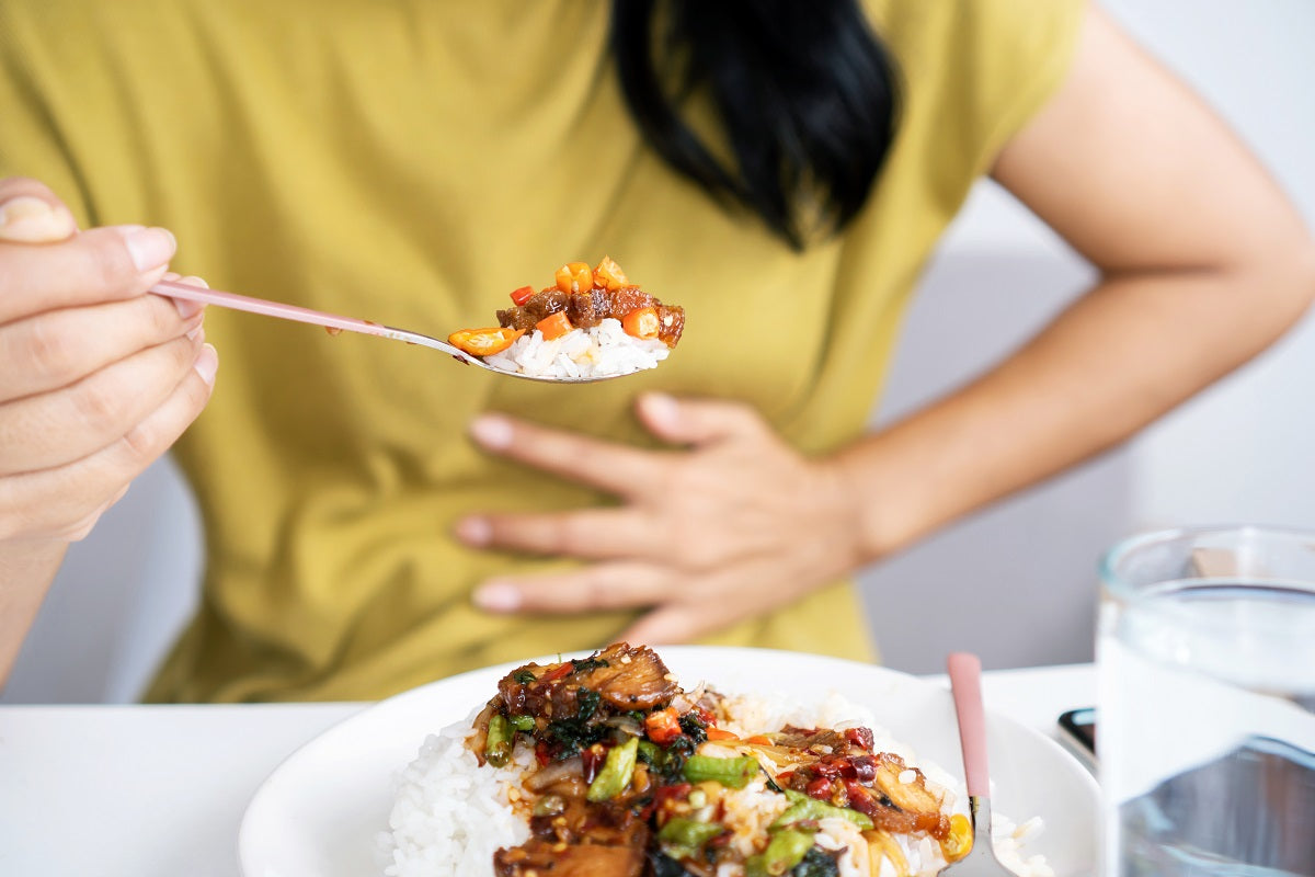 rau mit gelbem Oberteil hält sich beim Essen den Bauch, während sie einen Löffel mit Reis und einem scharfen Gericht zum Mund führt. Scharfes Essen kann Sodbrennen verursachen.