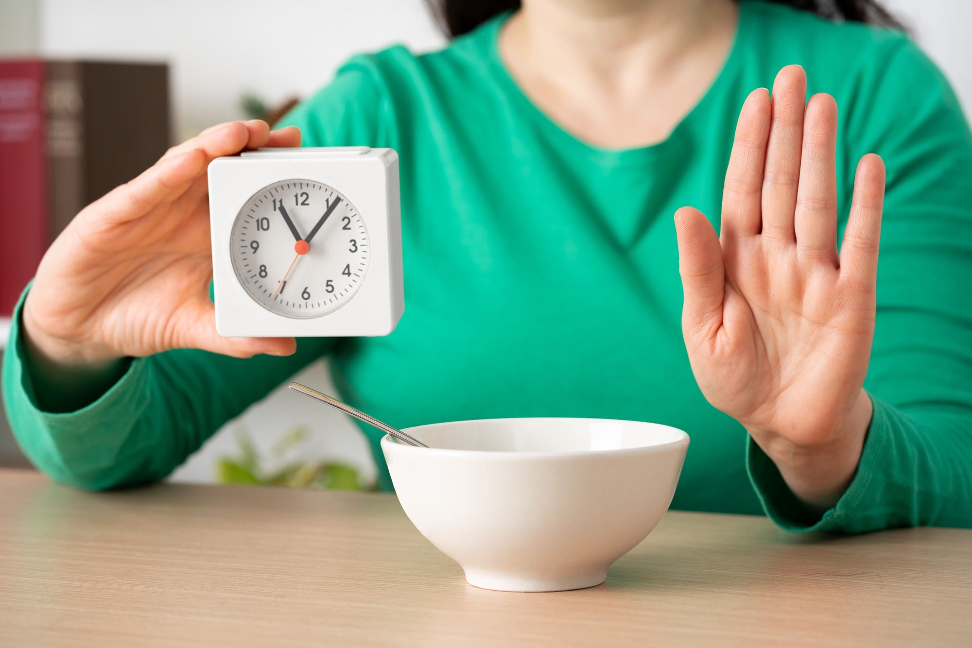 Frau in grünem Shirt zeigt auf eine Uhr und hebt die Hand zum Stoppzeichen – Symbolbild für achtsames Essen und bewusste Pausen beim Essen. Ideal für Beiträge über langsames Kauen, Verdauung und den Einfluss von Bitterstoffen auf das Sättigungsgefühl.