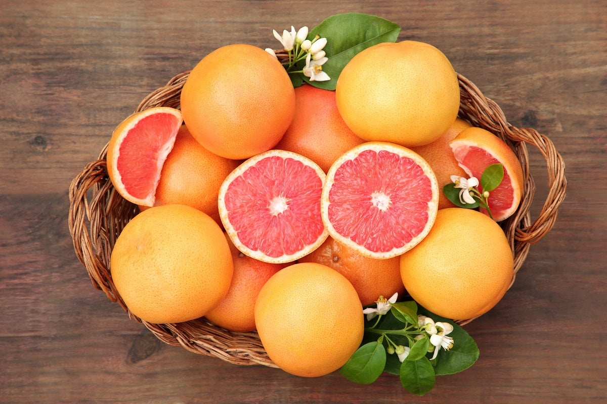 Ein geflochtener Korb voller rosa Grapefruits auf einem Holztisch, einige Früchte sind halbiert und zeigen das saftig-rote Fruchtfleisch; dekoriert mit grünen Blättern und kleinen weißen Blüten.