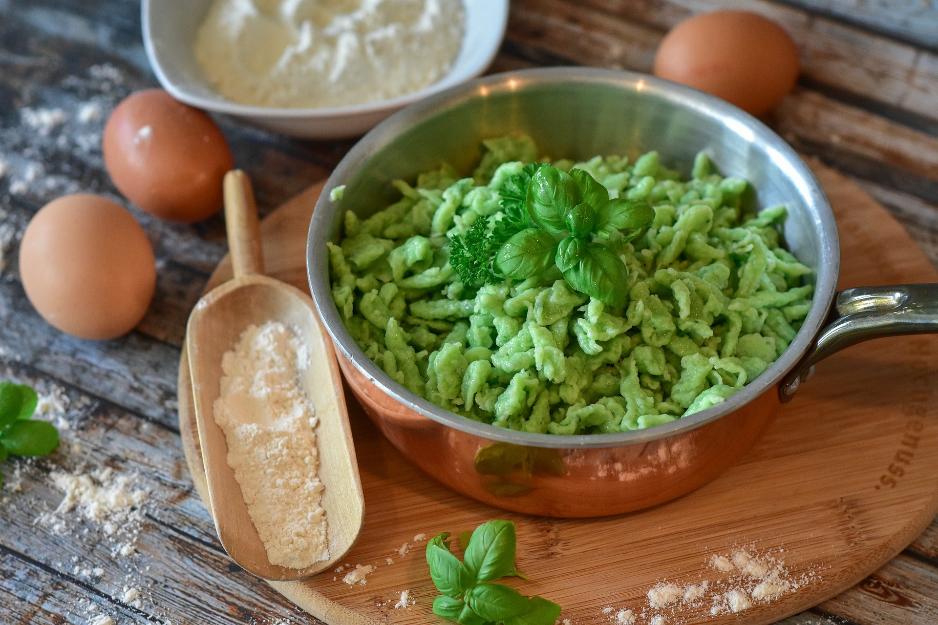 Brennnesselspätzle in einer Kupferpfanne auf einem Holzuntersetzer. Daneben ein Holzlöffel mit Mehl, eine Schüssel Mehl und drei Eier.