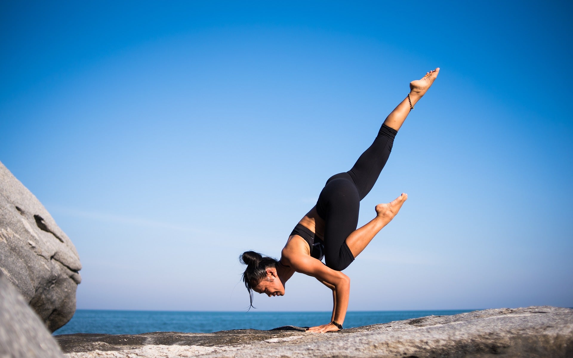 Eine sportliche Frau in schwarzer Trainingskleidung führt auf einem Felsen direkt am Meer eine anspruchsvolle Yoga-Balancehaltung aus. Ihr Körper ist kraftvoll angespannt, sie balanciert auf beiden Armen, während ein Bein nach oben und das andere nach hinten gestreckt ist. Der Hintergrund zeigt einen klaren blauen Himmel und das ruhige Meer