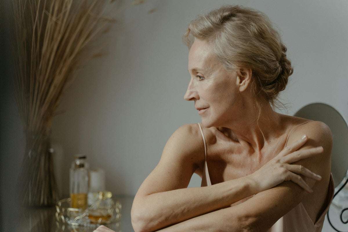 Frau im höheren Alter blickt zur Seite und lächelt leicht. Im Hintergrund sieht man Kosmetik, vielleicht Parfüm.