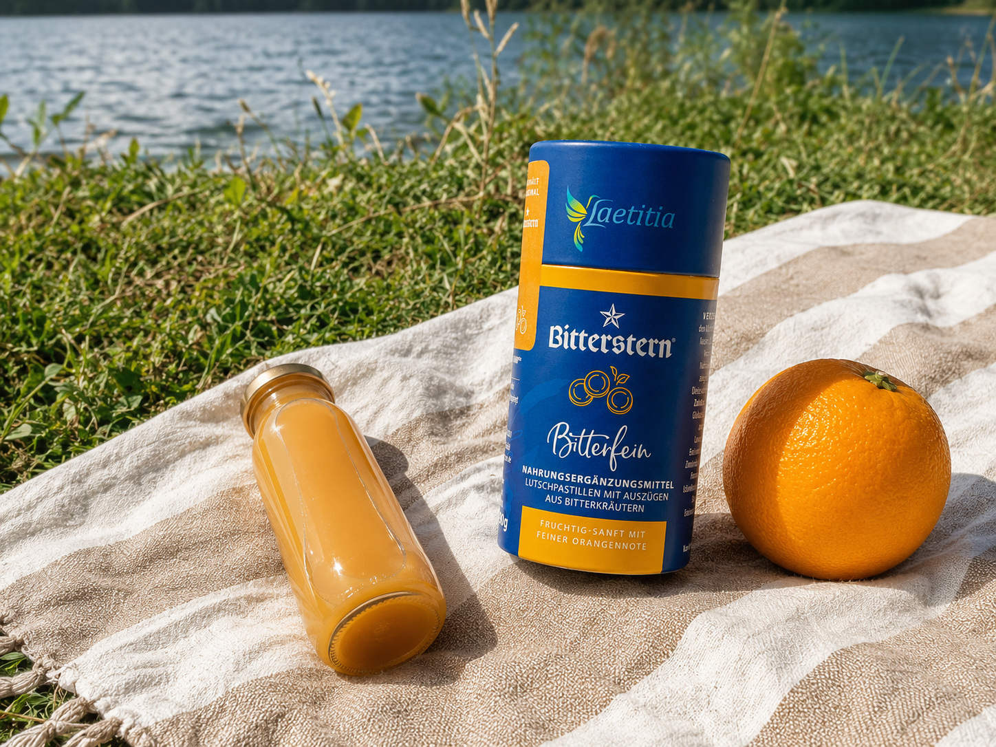 Eine Dose Bitterstern Pastillen, eine Orange und eine Flasche mit Fruchtsaft auf einer Picknickdecke am See
