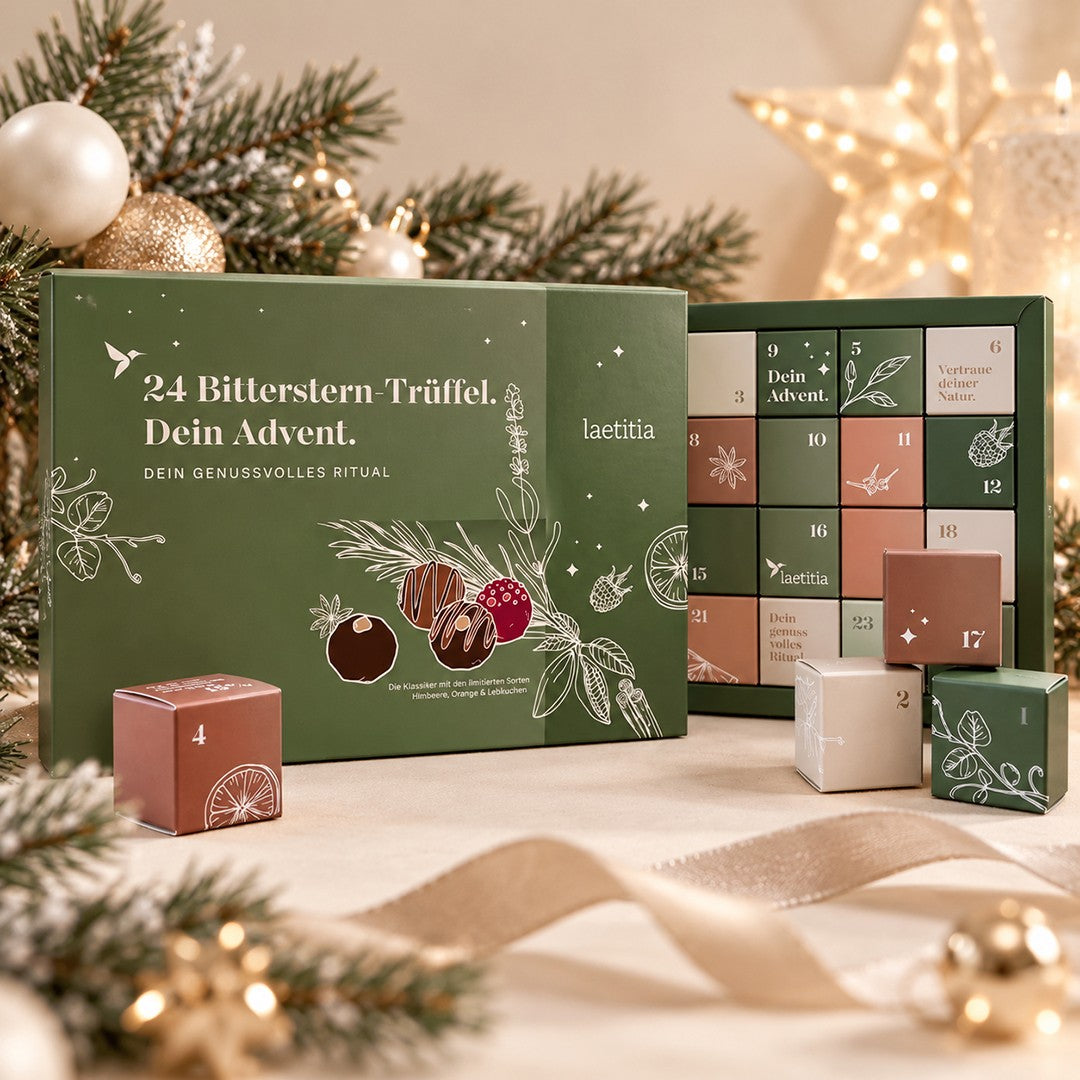 Geöffneter Bitterstern Trüffel Adventskalender mit 24 nummerierten Schachteln und Schokoladentrüffeln in den Sorten Himbeere, Orange und Lebkuchen, weihnachtlich dekoriert mit Tannenzweigen und Lichtern.