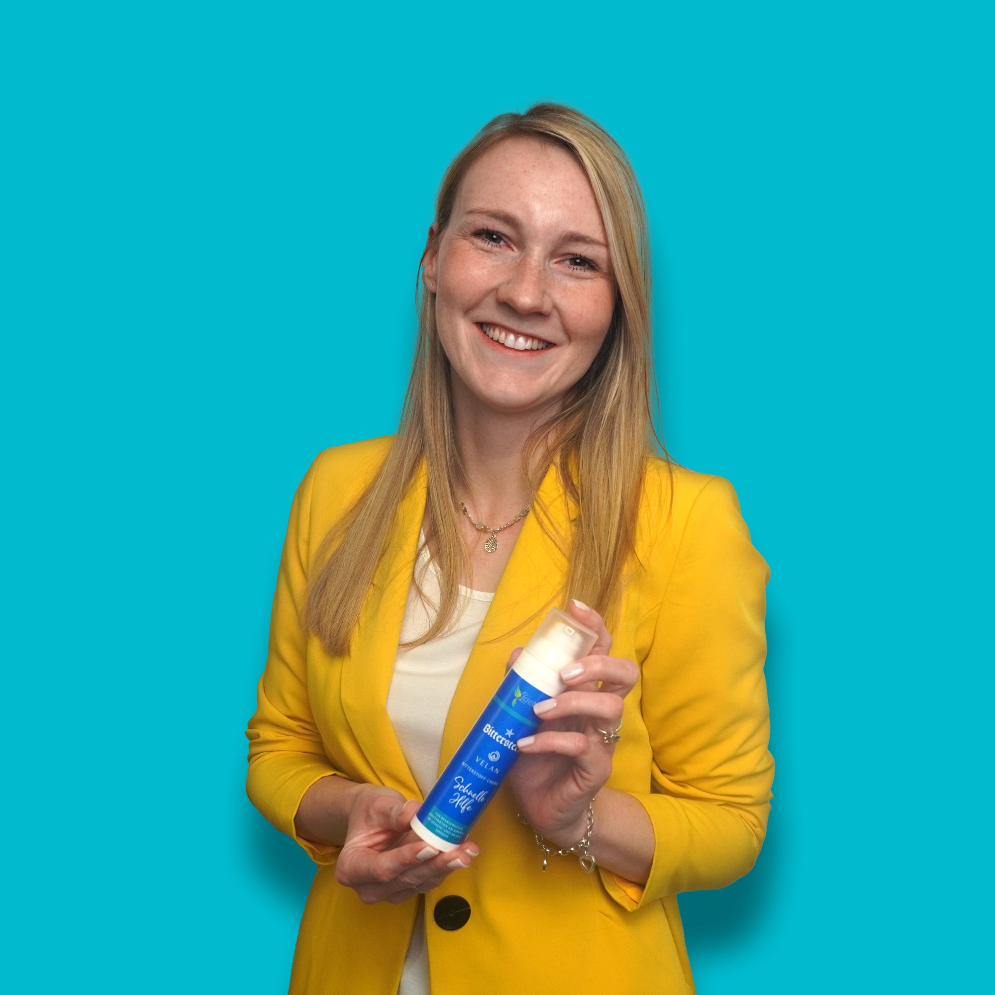 Eine lĂ€chelnde Frau mit blondem Haar in einem gelben Blazer steht vor einem tĂŒrkisblauen Hintergrund und hĂ€lt eine blaue Flasche in den HĂ€nden.