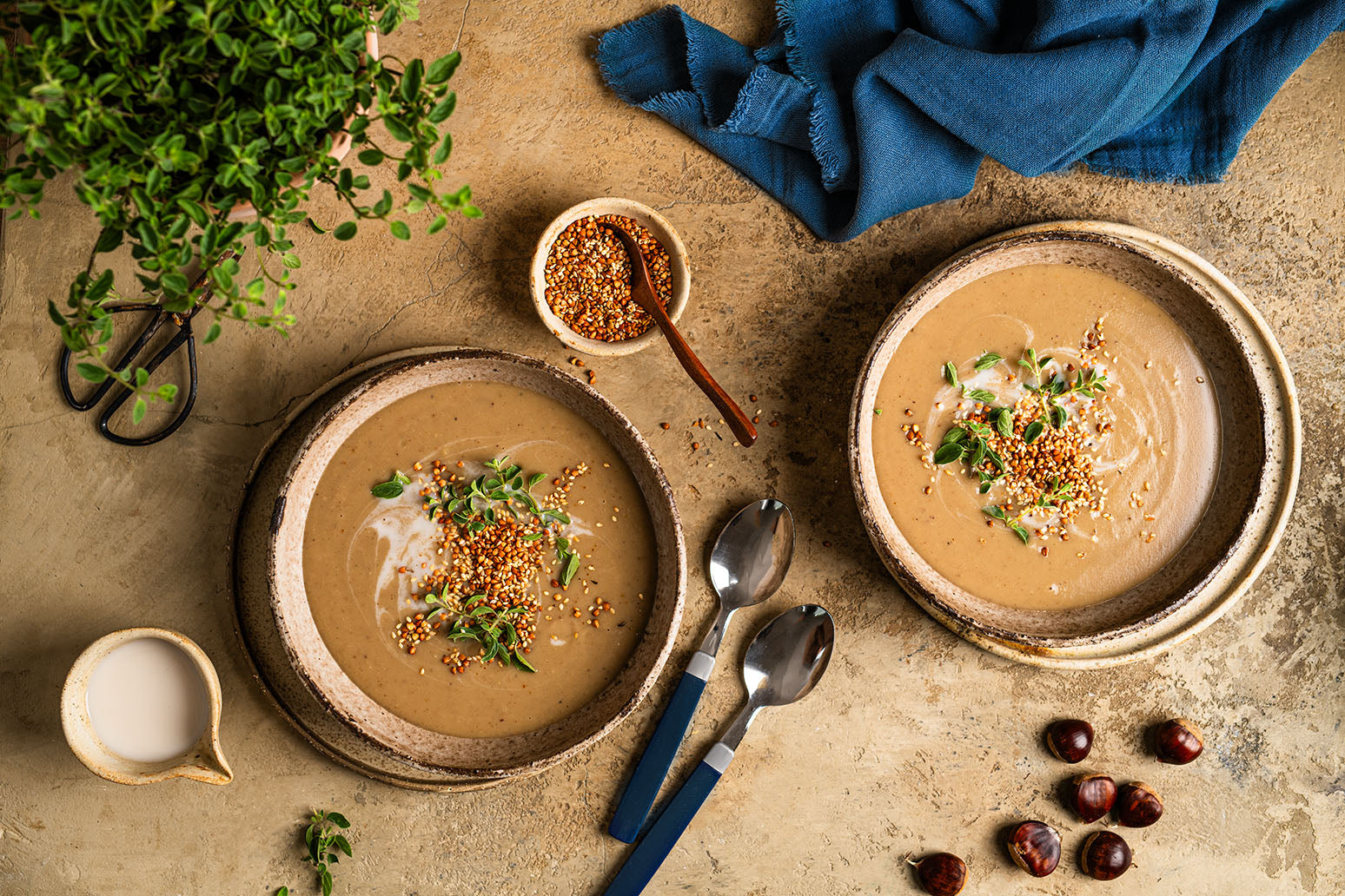 Basische Kastaniensuppe mit Buchweizen-Crunch in zwei beigen Tellern auf einem Steintisch - daneben eine blaue Serviette, eine kleine Schüssel Buchweizen, einzelne Kastanien, zwei Löffel und eine Kräuterpflanze. 