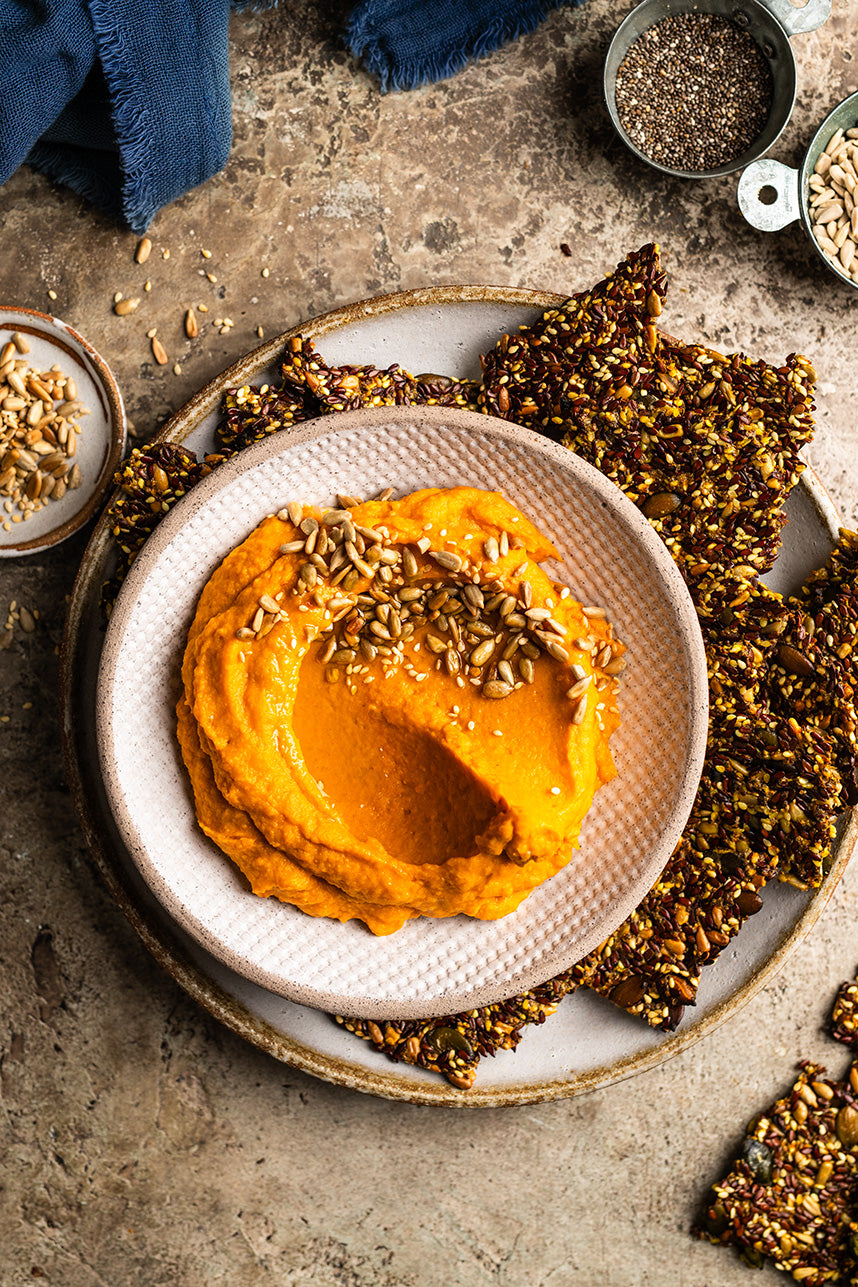 Süßkartoffel-Dip mit Crackern aus Körnern in einem Teller auf einem Holzbrett, außenherum Sonnenblumenkerne und Sesam in kleinen Schälchen, sowie ein blaues Geschirrtuch