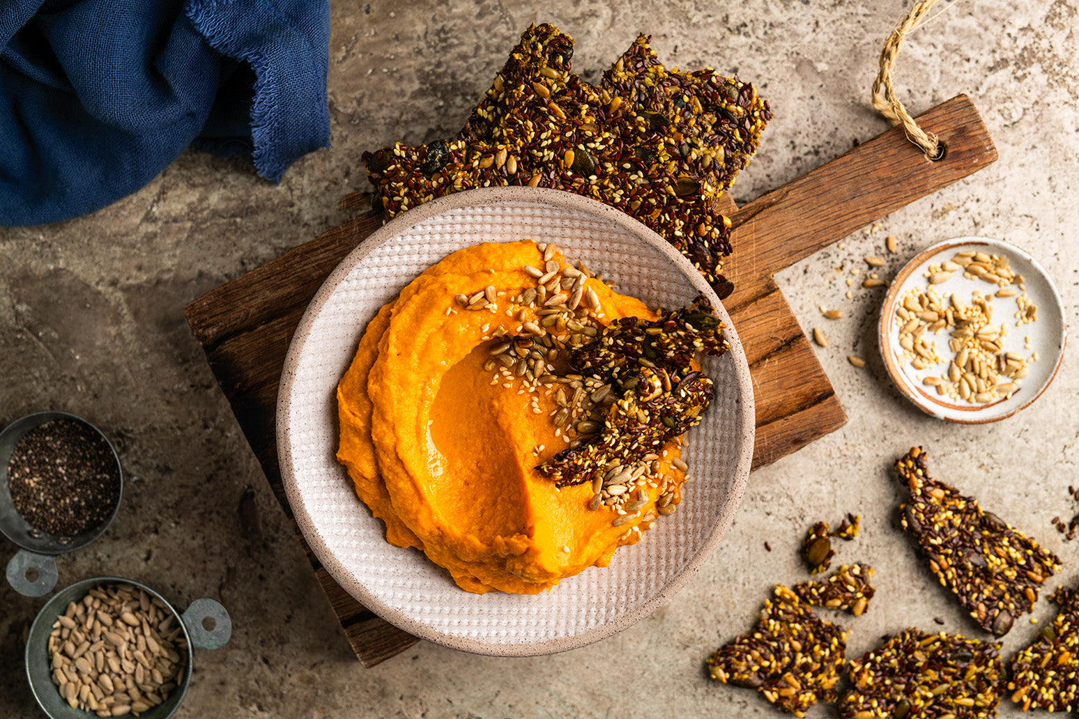 Süßkartoffel-Dip mit Crackern aus Körnern in einem Teller auf einem Holzbrett, außenherum Sonnenblumenkerne und Sesam in kleinen Schälchen