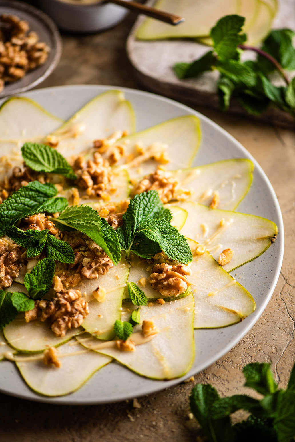 Birnen Carpaccio aus dünn geschnittenen Birnenscheiben, Walnüssen und Minze kreisförmig auf einem Teller angerichtet.