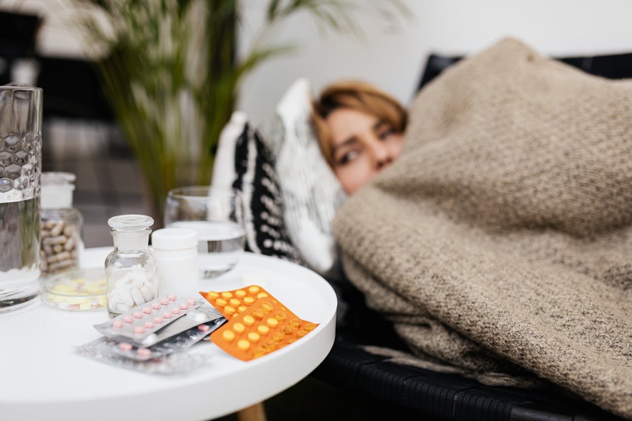 Frau liegt mit einem Infekt unter der Decke, daneben ein Nachttisch mit Tabletten