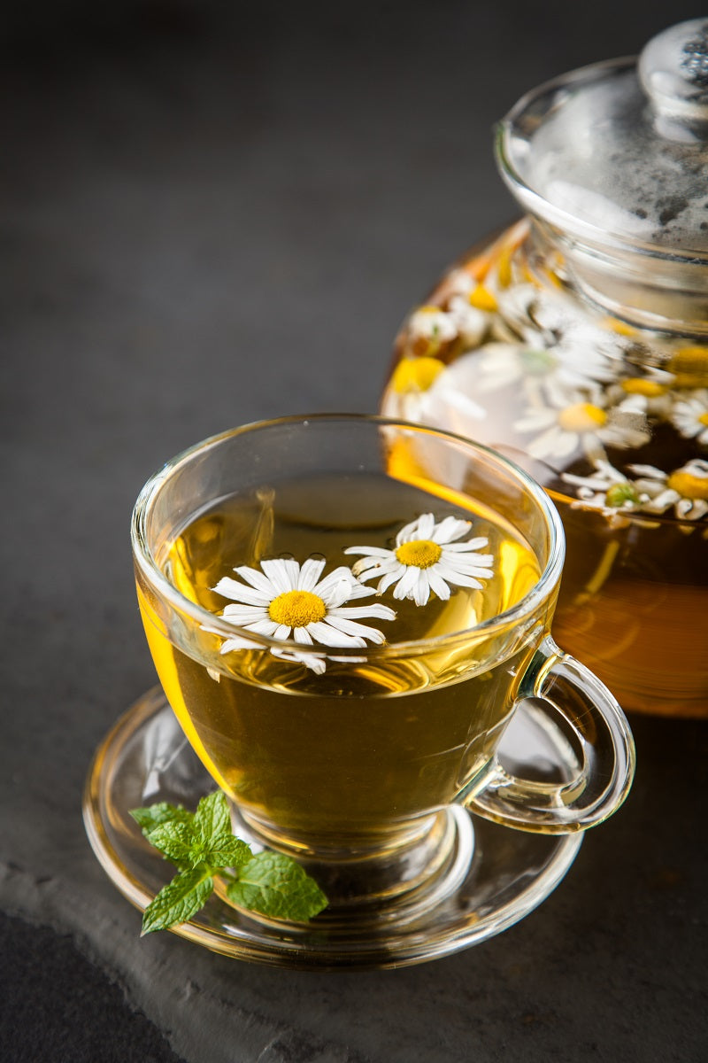 Eine Tasse Tee mit Gänseblümchen vor einem dunklen Hintergrund - im Anschnitt ist eine Teekanne zu erkennen