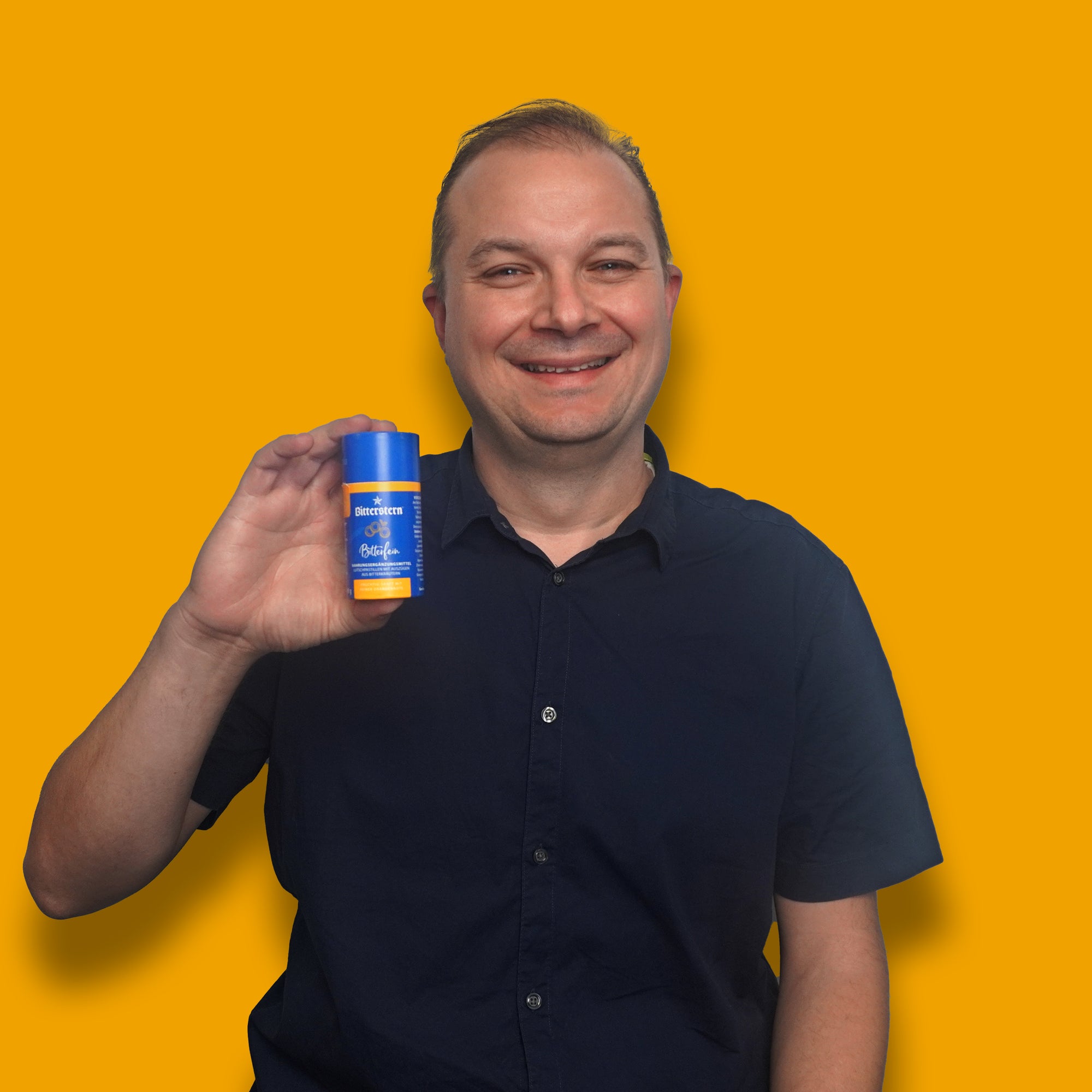 Mann im dunkelblauen Hemd lÀchelt in die Kamera und hÀlt eine Dose Bitterstern Pastillen vor orangefarbenem Hintergrund in der Hand.