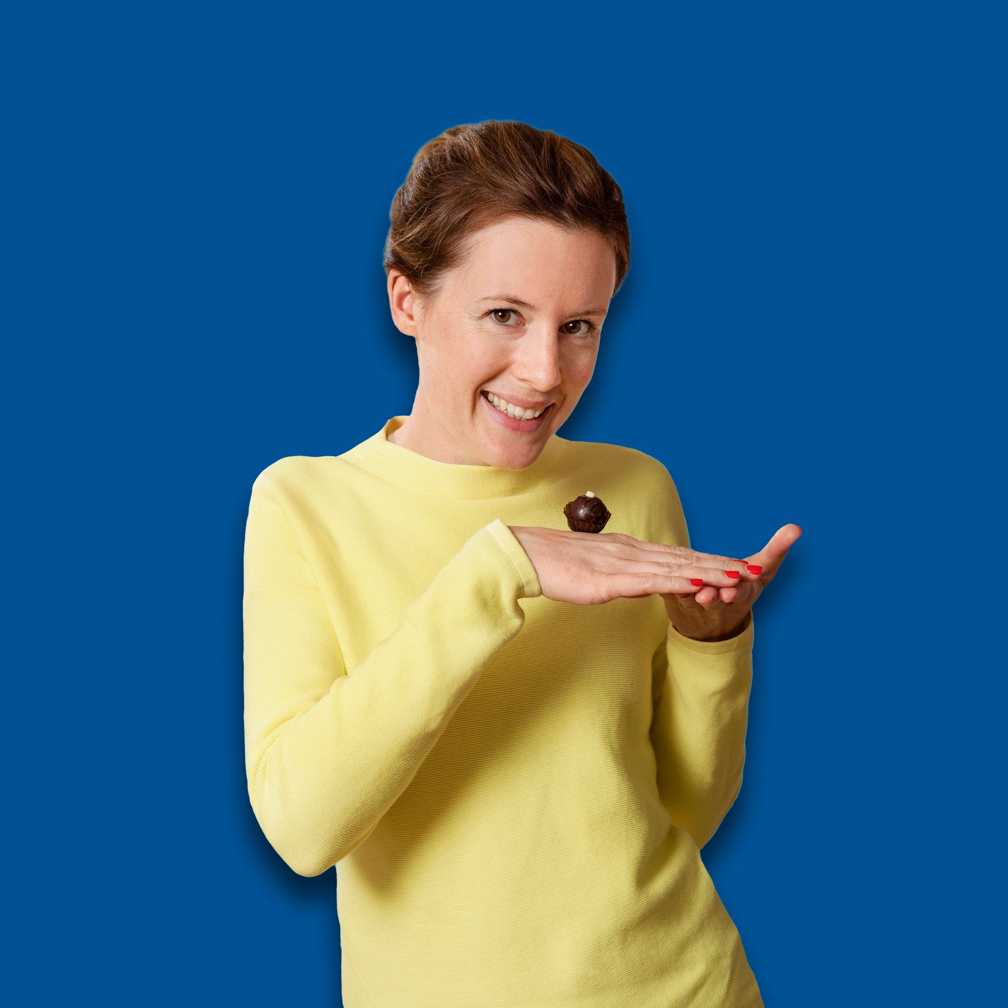 Frau im gelben Pullover lĂ€chelt, balanciert einen Bitterstern SchokoladentrĂŒffel auf dem HandrĂŒcken. Blauer Hintergrund