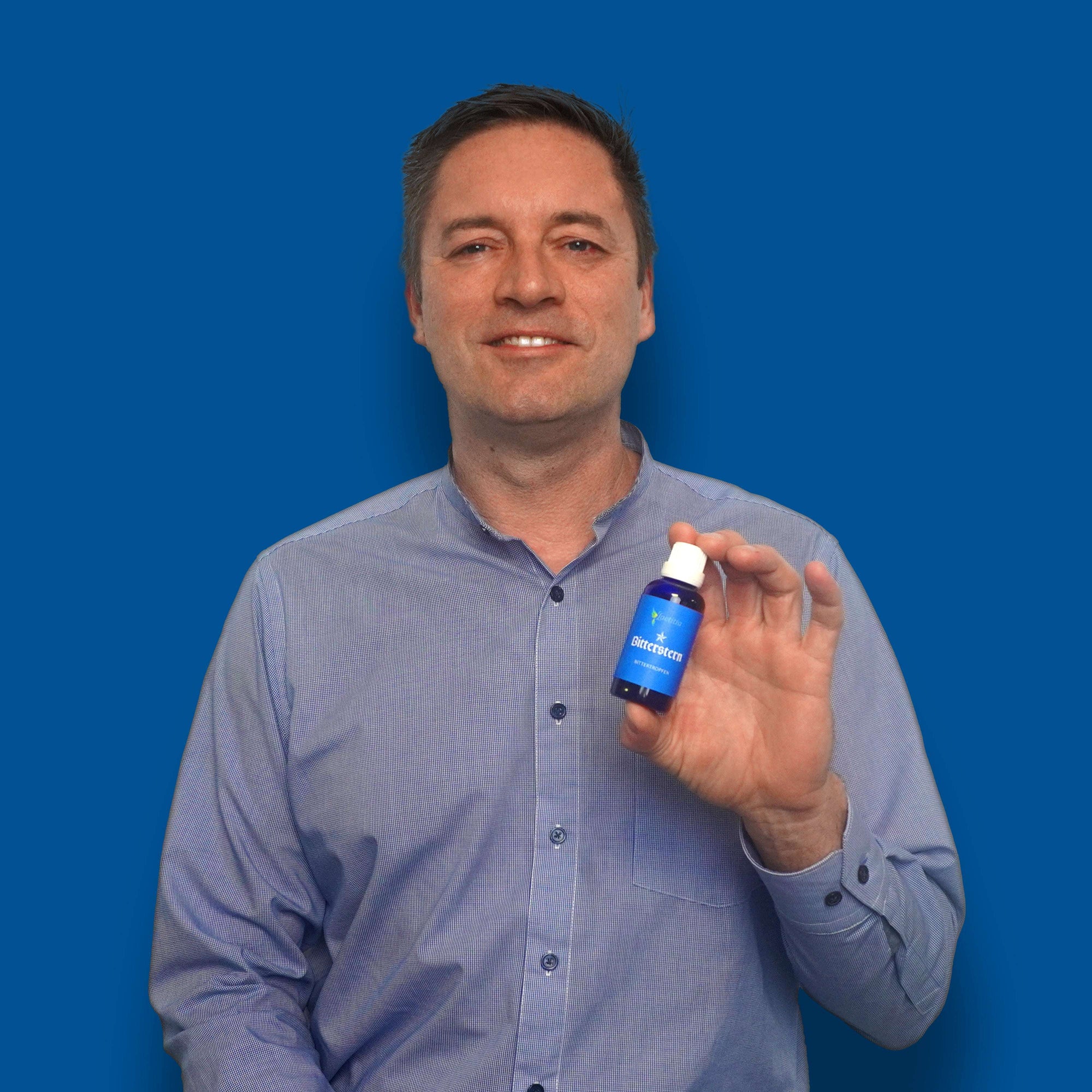 Mann im karierten Hemd hÀlt Bitterstern Bittertropfen in der Hand, blauer Hintergrund.