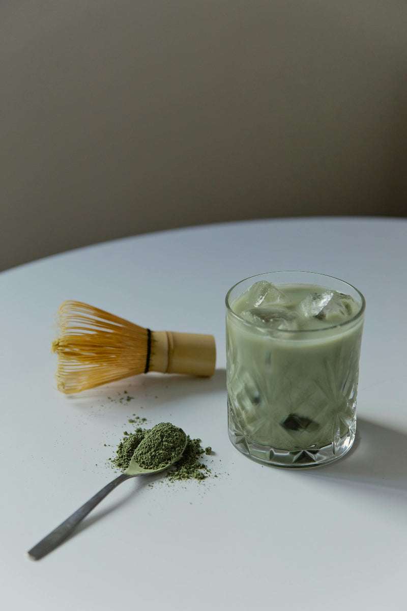 Ein Teelöffel mit Matchapulver darauf, daneben ein Matchabesen und ein Glas gefüllt mit Eiswürfeln und Matcha Latte auf einem weißen Tisch