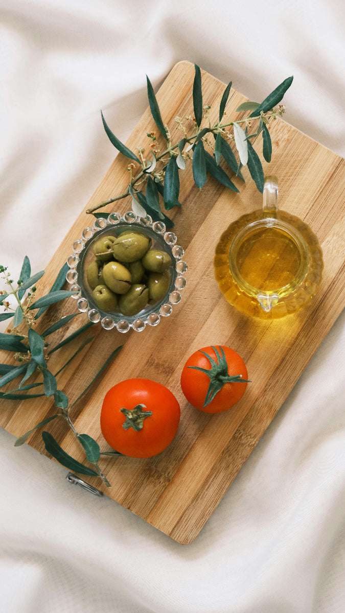 Holzbrett mit typisch mediterranen Zutaten, wie Olivenöl, Oliven und Tomaten auf einem weißen Tischtuch