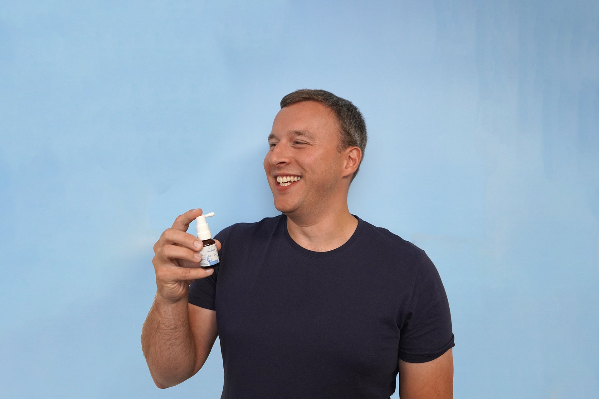 Mann im dunkelblauen T-Shirt vor einem hellblauen Hintergrund hält Bitterstern Bitter-Spray in der Hand. Lächelt. 