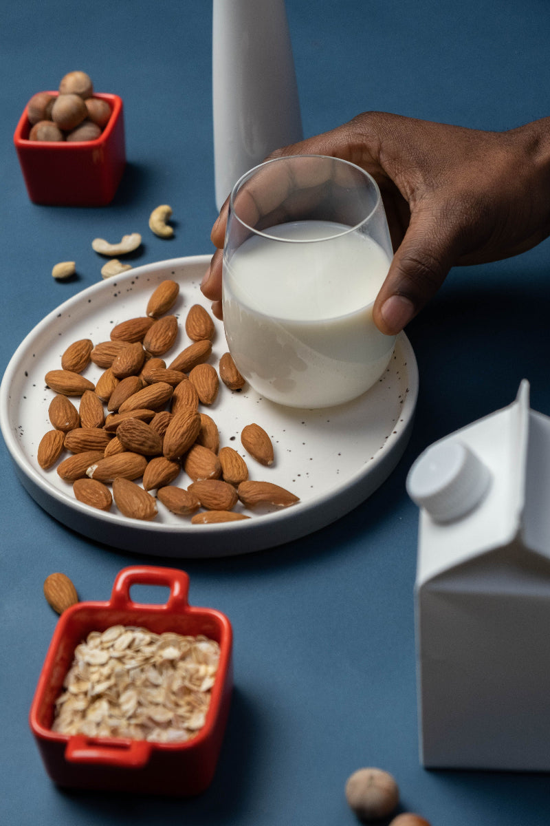 Ein Glas Milch auf einem kleinen Teller, daneben ein paar Mandeln und neben dem Teller noch 2 kleine Schälchen mit weiteren Lebensmitteln, wie Haselnüssen und Haferflocken auf einem blauen Tisch
