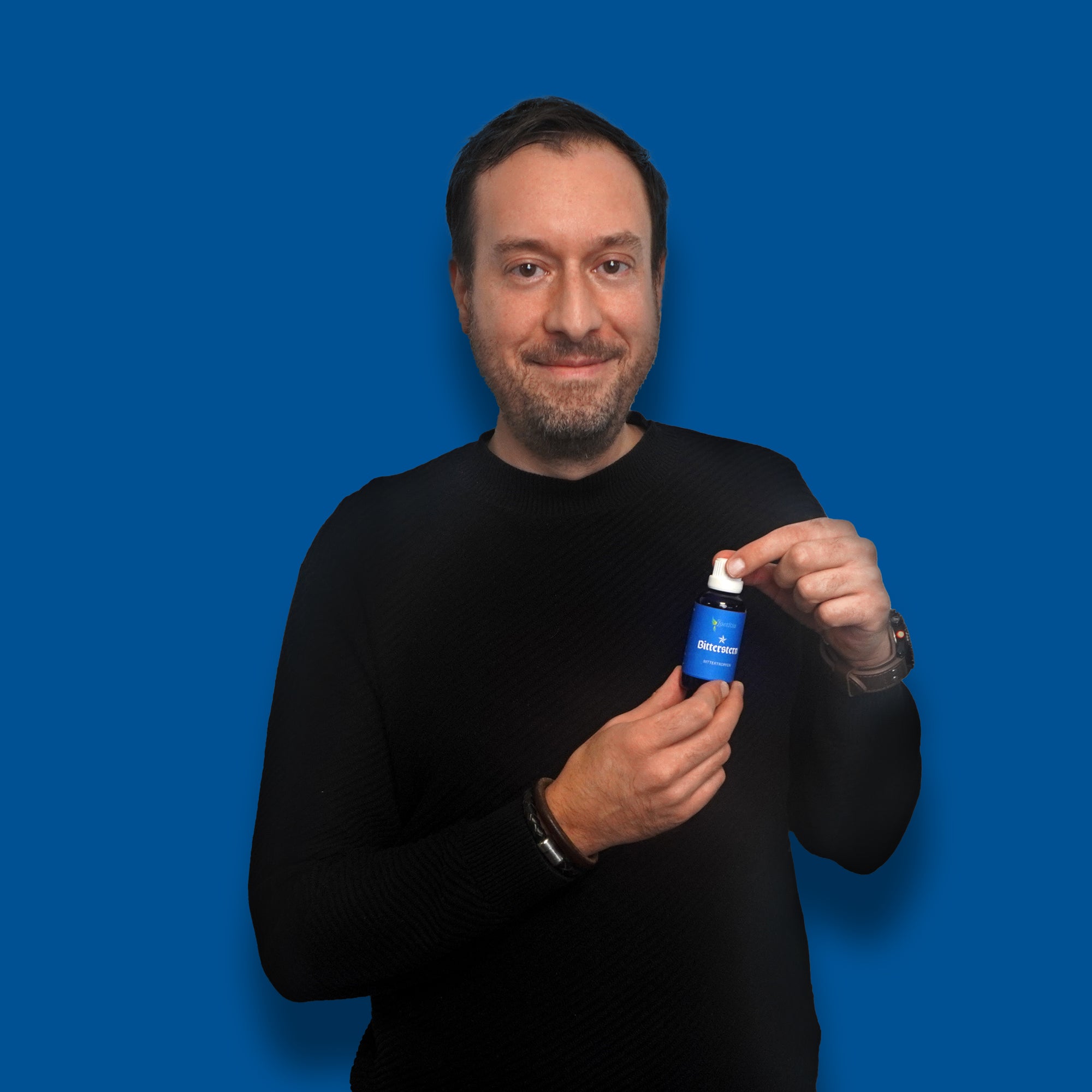 Ein Mann mit dunklem Haar und Bart trĂ€gt einen schwarzen Pullover und steht vor einem blauen Hintergrund. Er lĂ€chelt und hĂ€lt eine blaue Flasche Bitterstern Tropfen mit weiĂem Deckel in beiden HĂ€nden.